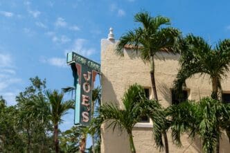 The iconic vertical sign for Joe's Stone Crab restaurant in Miami Beach, surrounded by lush green palm trees against a blue sky.