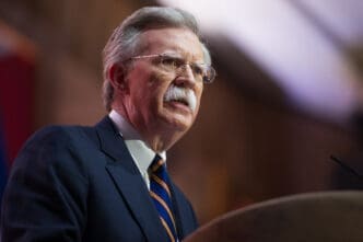 Close-up portrait of John Bolton, wearing a dark suit and striped tie, speaking into a microphone at a podium.