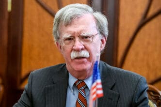 Close-up portrait of John Bolton, former US National Security Advisor, wearing a suit and glasses, with a small American flag in the foreground.