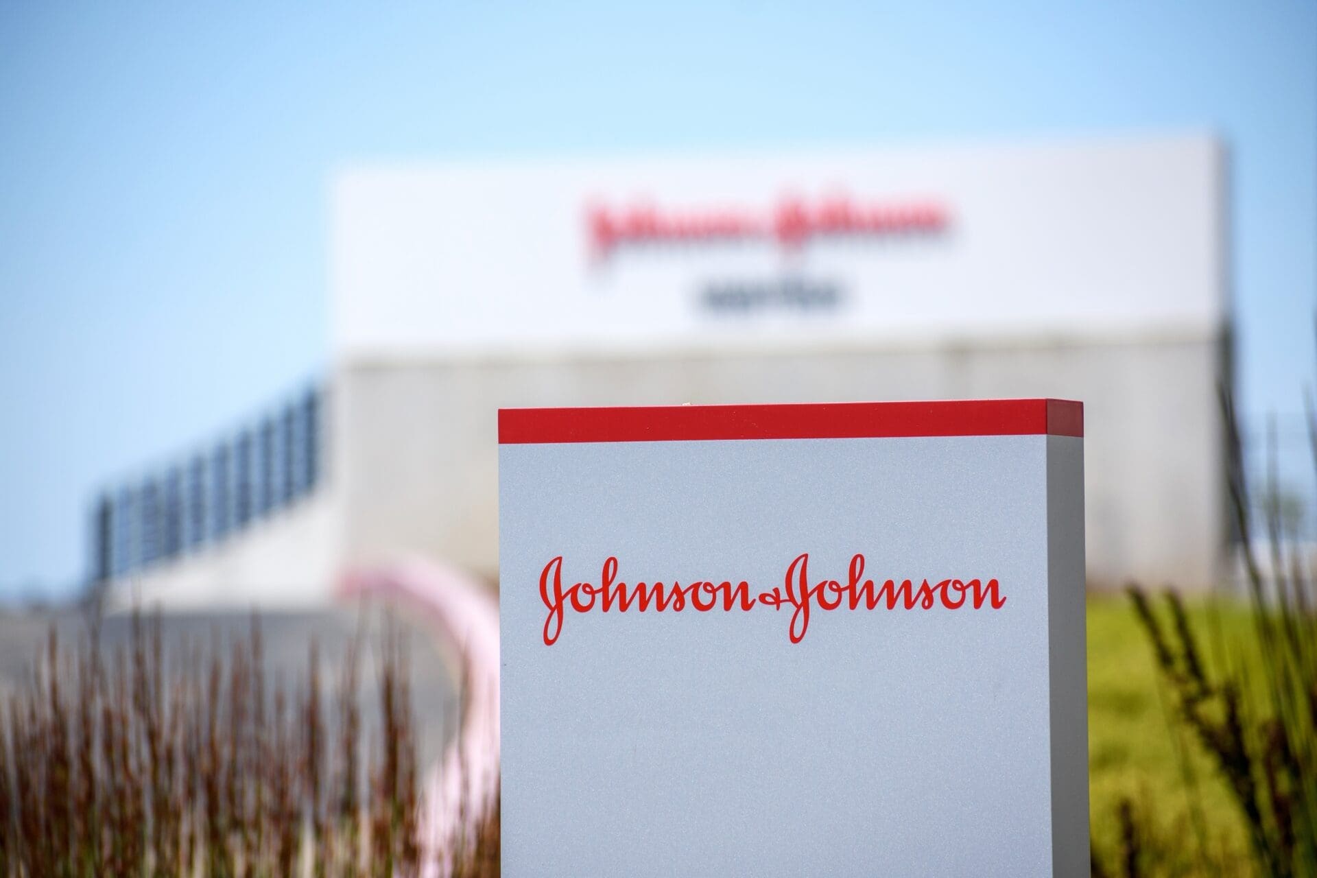 A close-up of a silver sign with the red Johnson & Johnson script logo in front of a blurred corporate building.