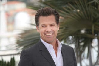 Actor Josh Brolin smiling widely, wearing a dark gray suit jacket and open-collared white shirt outdoors