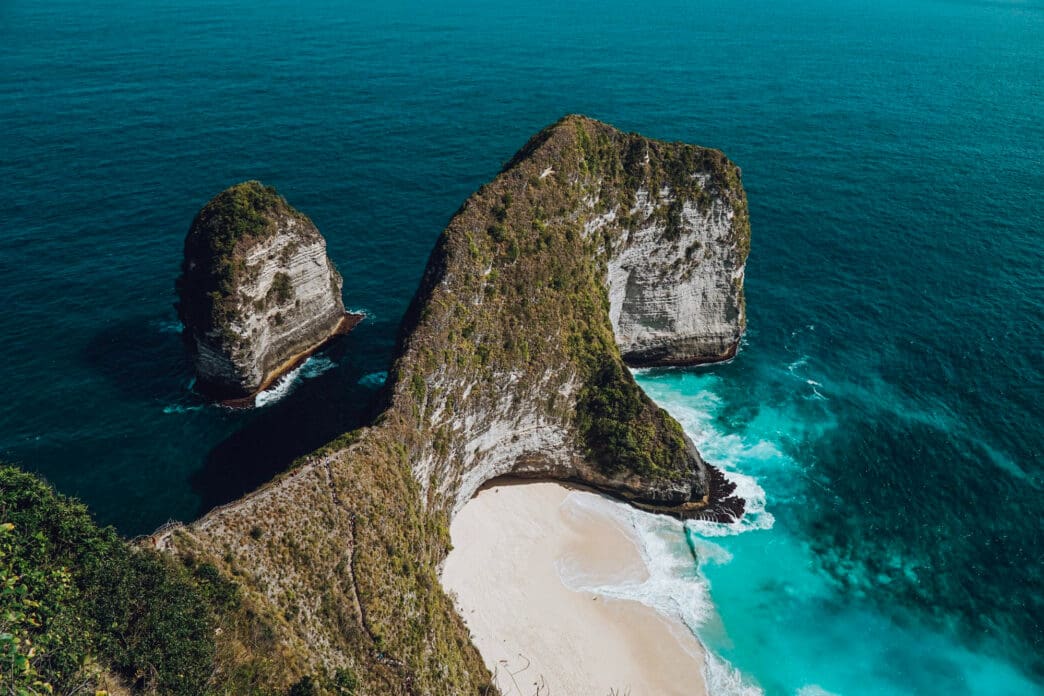 A massive T-Rex shaped cliff towering over a secluded white sand beach and turquoise ocean in Bali.