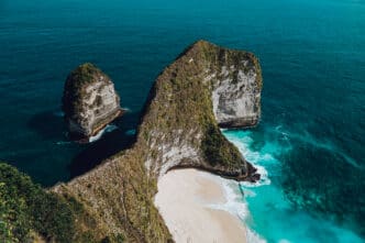 A massive T-Rex shaped cliff towering over a secluded white sand beach and turquoise ocean in Bali.