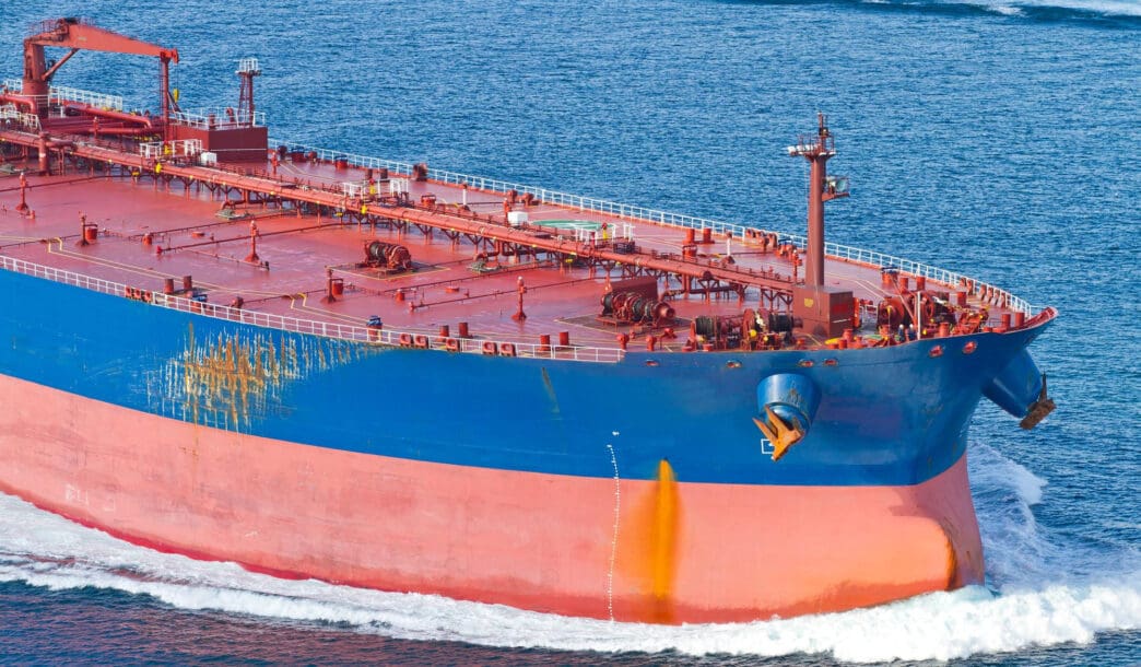 Close-up view of the bow of a massive oil tanker ship painted red and blue, cutting through deep blue ocean water.
