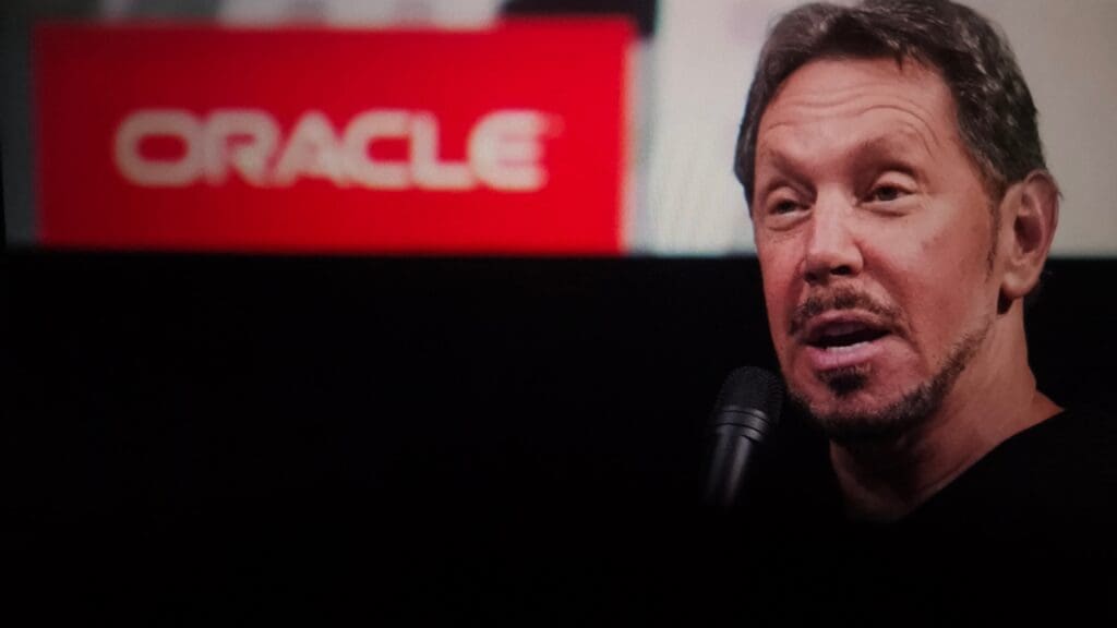 Close-up of Oracle co-founder Larry Ellison speaking into a microphone with the bright red "ORACLE" logo visible in the blurred background