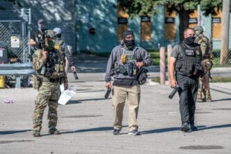 Three heavily armed law enforcement officers, including one in military camouflage and one wearing an "ICE Police" vest, stand guard outside a facility.