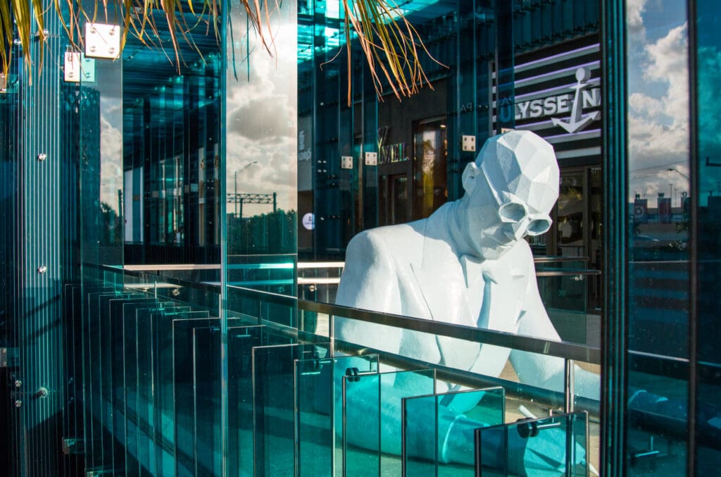 Polygonal sculpture of Le Corbusier in a suit and glasses reflected in a modern blue-tinted glass facade