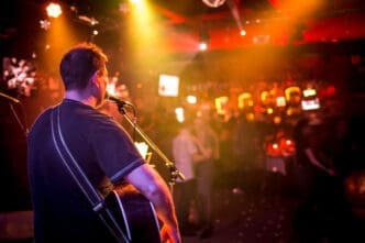 Musician with an acoustic guitar and a black shirt performs on stage under warm spotlights facing a blurry crowd in a bar.