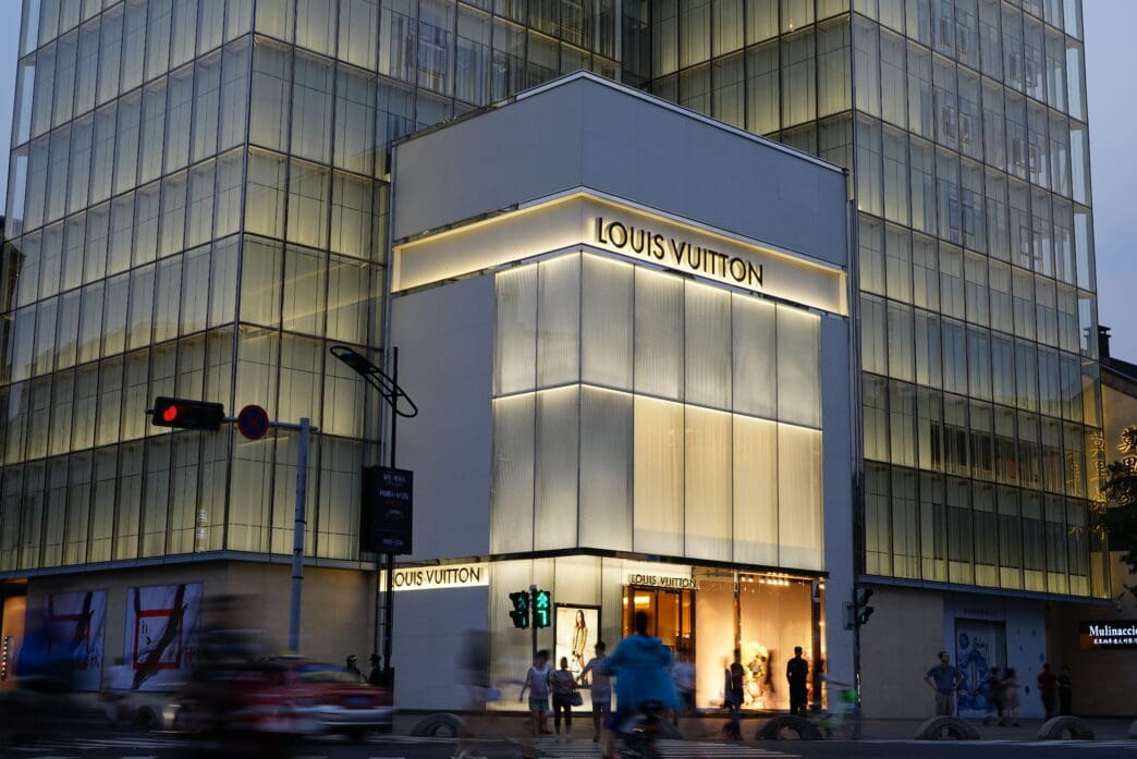 Evening view of a brightly lit, modern Louis Vuitton flagship store with a glass tower backdrop in a Chinese city.