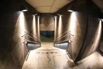 A fisheye view looking down a well-lit, modern concrete staircase leading into a dark, subterranean passageway or bunker.
