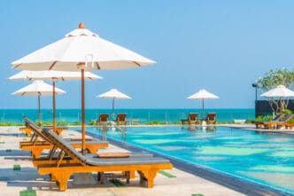 Row of empty wooden lounge chairs and white umbrellas by an infinity pool overlooking the bright blue ocean.