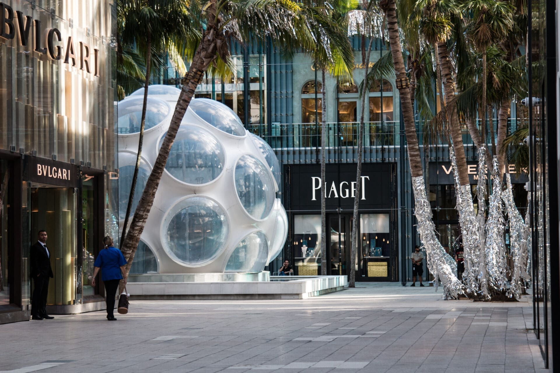 Outdoor view of Miami Design District showing high-end stores like Bulgari and Piaget, palm trees, and a futuristic bubble-like structure.
