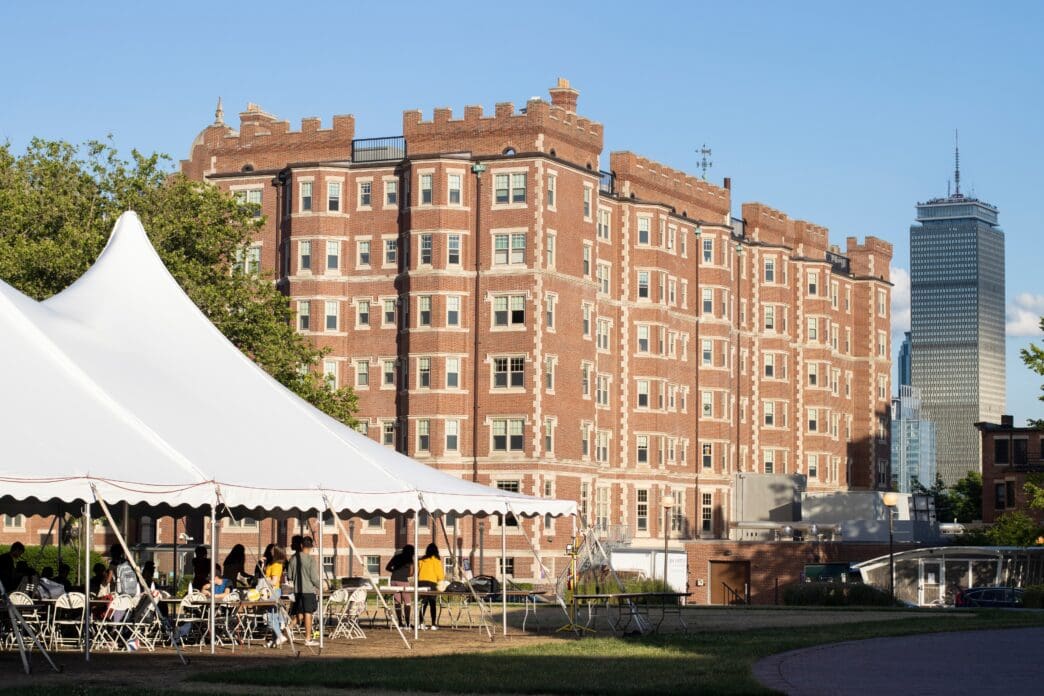 Fariborz Maseeh Hall (MIT Building W1), a large, fortress-like brick dormitory, stands behind a white tent with students, with the Prudential Tower visible in the distance.