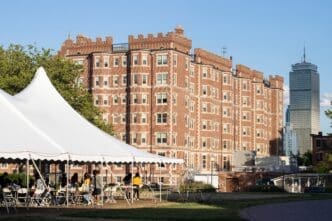 Fariborz Maseeh Hall (MIT Building W1), a large, fortress-like brick dormitory, stands behind a white tent with students, with the Prudential Tower visible in the distance.