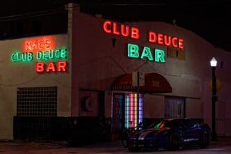 A blue sports car parked outside the neon-lit Mac's Club Deuce bar at night.