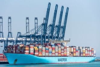 Massive light blue Maersk container ship fully loaded with stacked containers is docked beneath towering gantry cranes at a shipping port.