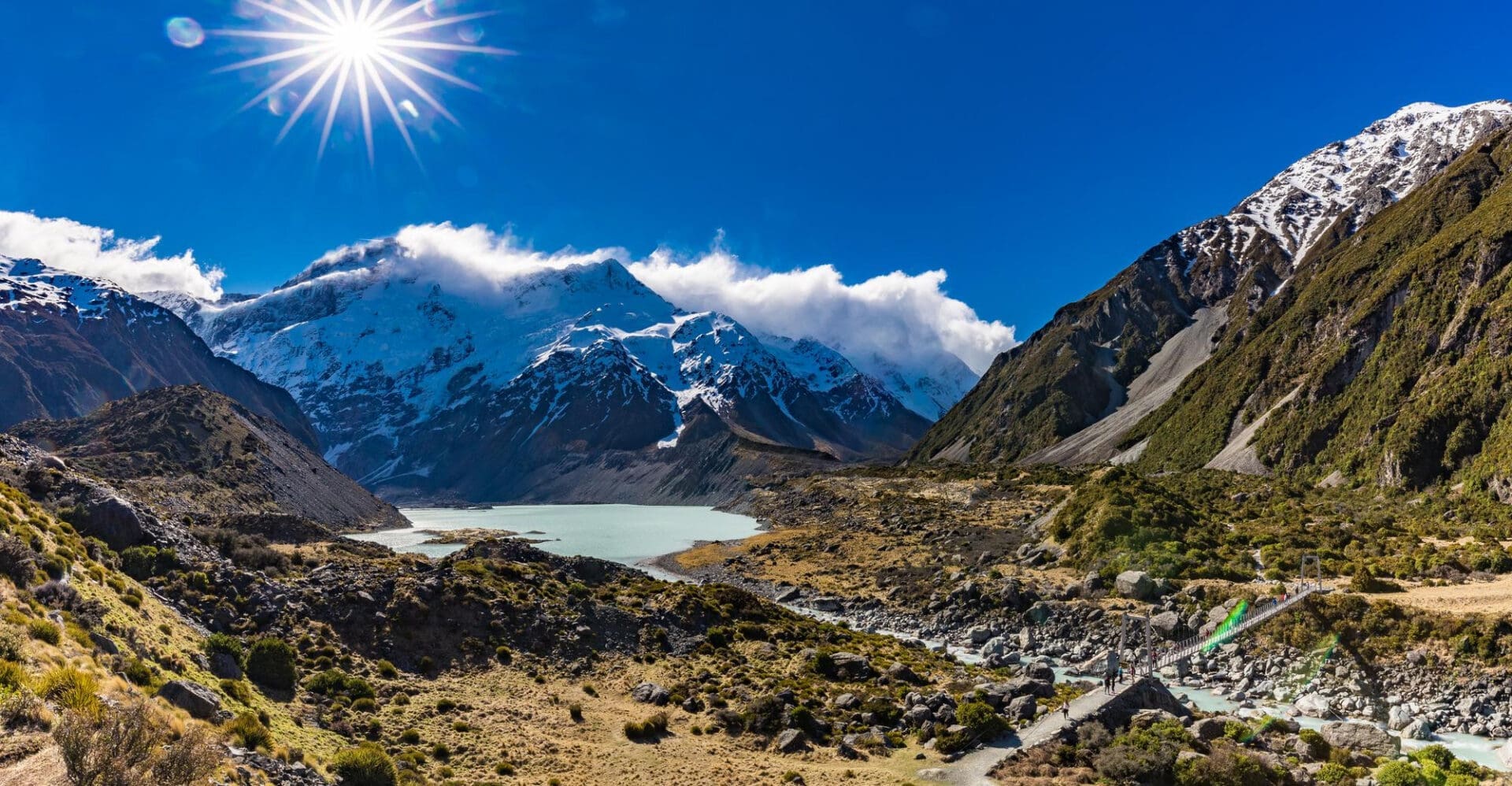 Sunlit panoramic view of the Hooker Valley Track in Aoraki National Park, showing snow-capped mountains, a glacial lake, and a river with a swing bridge.