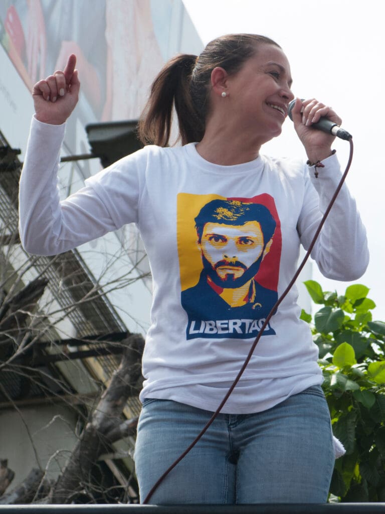 María Corina Machado speaking into a microphone at a rally, wearing a shirt with a portrait of a bearded man and the word "LIBERTAD"