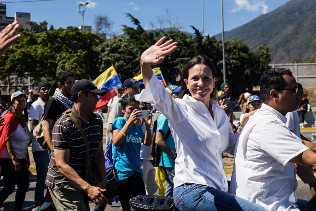 Venezuelan opposition leader María Corina Machado smiling and waving to a crowd of supporters from the back of a motorcycle.