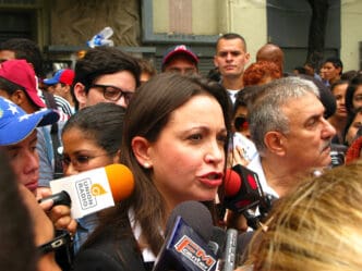 Opposition leader Maria Corina Machado speaking intensely into a cluster of reporters' microphones during a crowded street protest