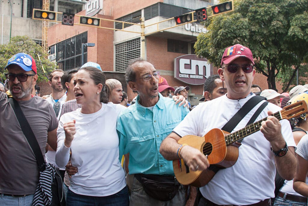 Maria Corina Machado and two men singing passionately in a crowded street protest, with one man playing a Venezuelan cuatro