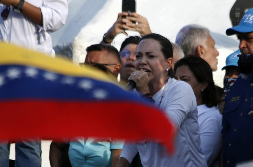 Venezuelan opposition leader María Corina Machado speaking passionately into a microphone at a rally, partially obscured by the Venezuelan flag
