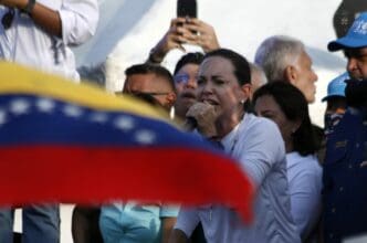 Venezuelan opposition leader María Corina Machado speaking passionately into a microphone at a rally, partially obscured by the Venezuelan flag