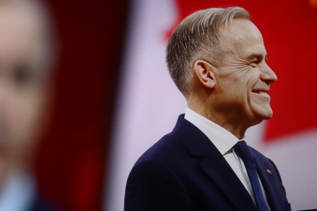 Profile close-up of a smiling Mark Carney in a suit against a blurred red and white background that includes a second image of him.