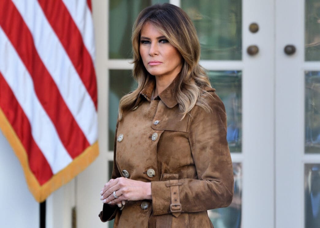 First Lady Melania Trump stands in a brown coat in the White House Rose Garden.