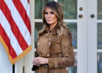 First Lady Melania Trump stands in a brown coat in the White House Rose Garden.