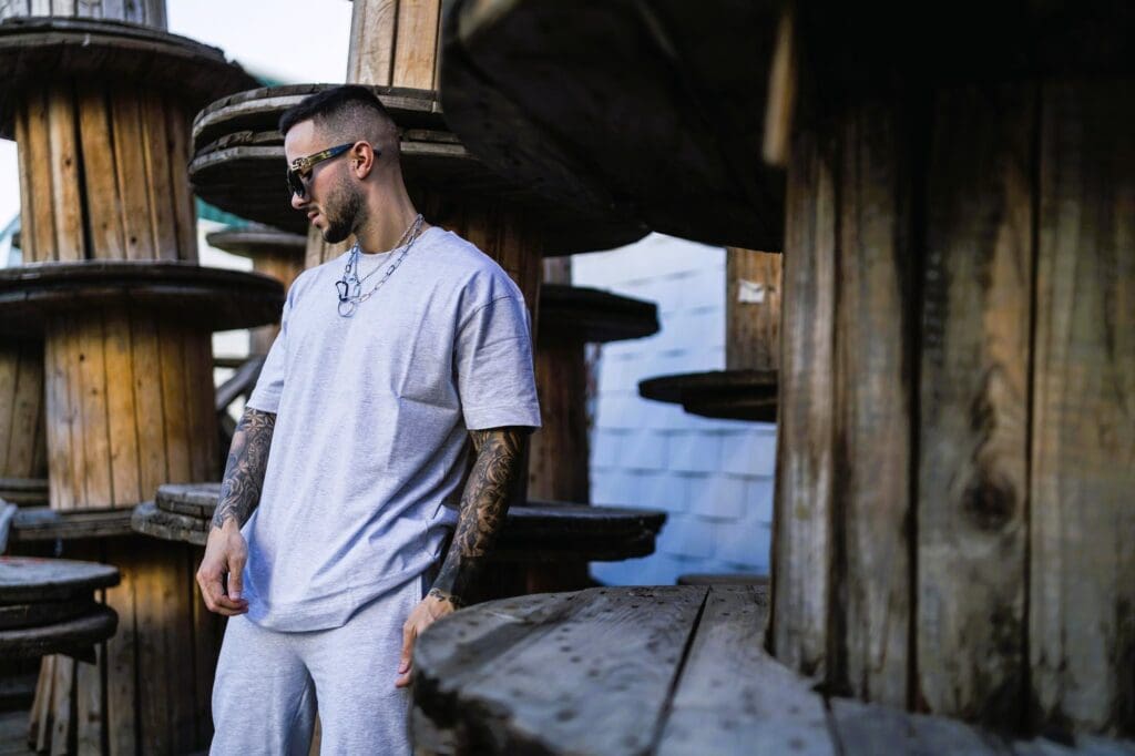 Stylish man with tattoos wearing a gray oversized t-shirt, matching sweatpants, and sunglasses posing by large wooden spools.
