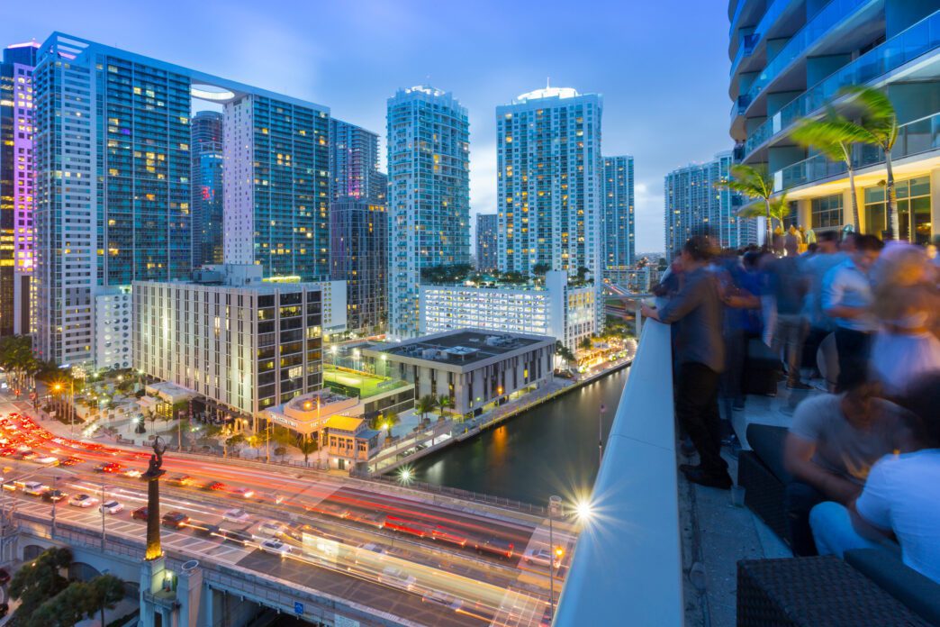 A rooftop bar in Miami buzzes with people overlooking the Brickell Avenue bridge and city skyline at dusk.