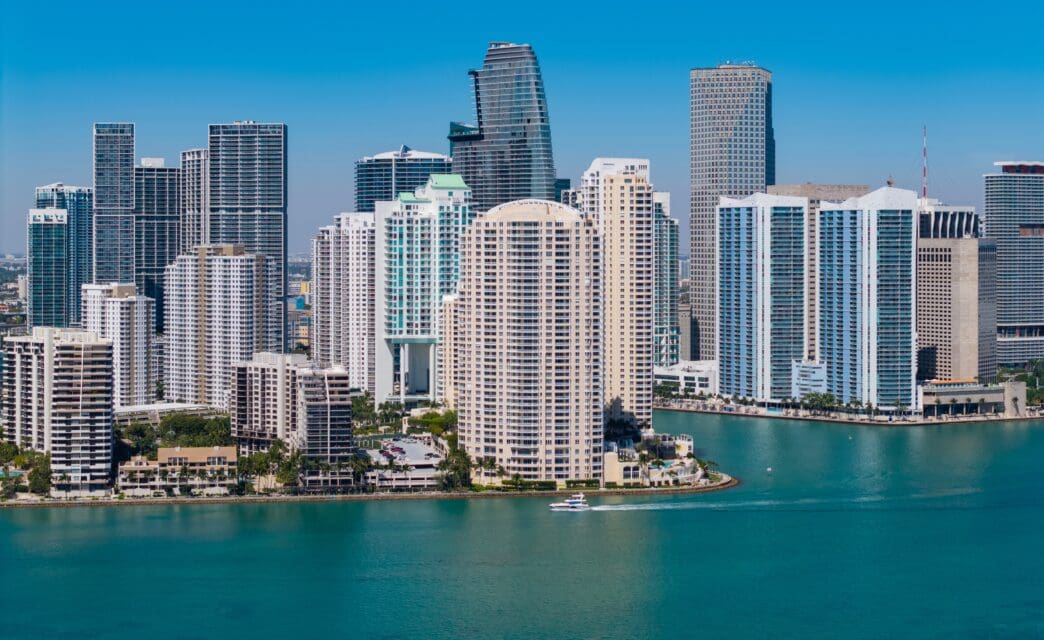 A boat moves across the turquoise water in front of the Brickell Key and downtown Miami skyline on a sunny day.