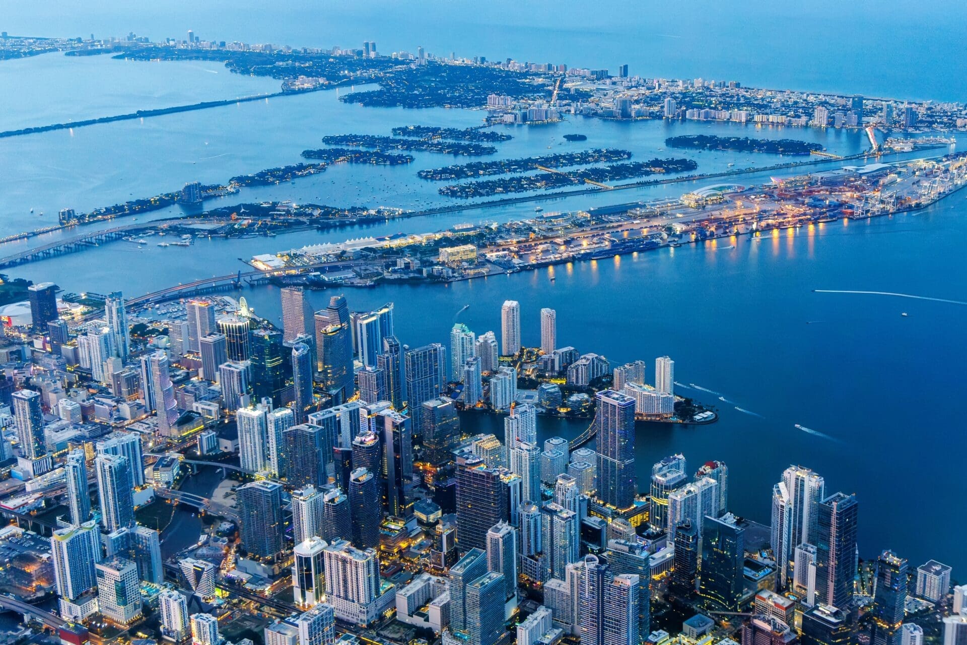 High aerial view of the illuminated Miami skyline at twilight, showing Downtown, Brickell, and Miami Beach across the bay.