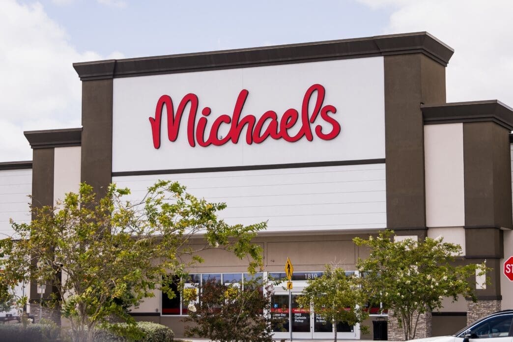 Exterior view of a Michaels craft store building featuring its logo in red script letters on a white and brown facade.