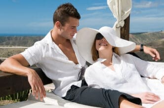 Attractive couple in white shirts relaxing on an outdoor lounge bed in a sunny, arid Mediterranean setting.