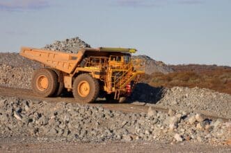 A large, yellow haul truck drives on a dirt road in a rocky open-pit mine, with piles of ore.