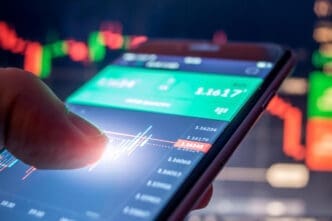 A person's finger touches a smartphone screen displaying a trading app with a live stock or forex candlestick chart and glowing data.