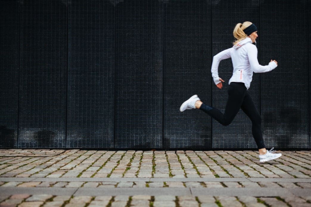 Profile of a young woman running outdoors in black leggings and a white jacket along a cobbled sidewalk.