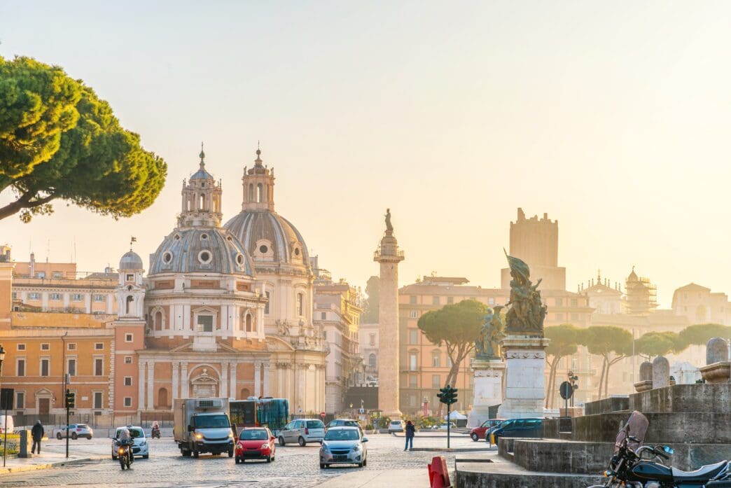 A morning view of Rome's Trajan Forum, showing historic churches, Trajan's Column, and street traffic.