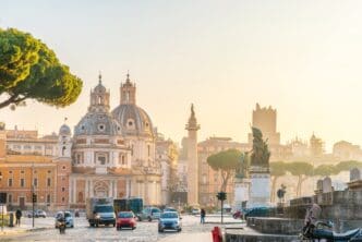 A morning view of Rome's Trajan Forum, showing historic churches, Trajan's Column, and street traffic.