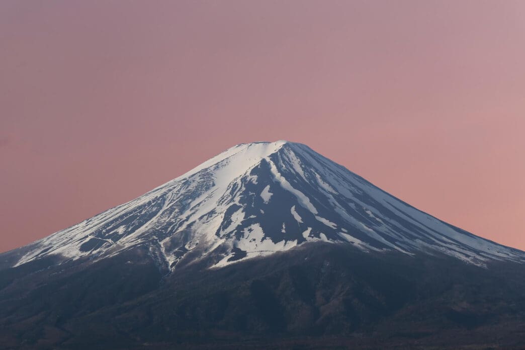 A close-up of the snow-capped peak of Mount Fuji against a pink sky.
