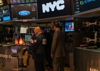 Traders and executives standing on the NYSE floor, dominated by screens showing stock prices and the Ford Motor Company logo