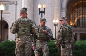 Three National Guard soldiers in uniform stand in conversation outside a building with ornate architecture and lampposts.