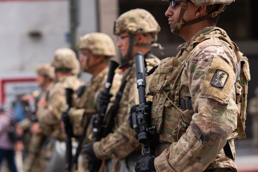 National Guard soldiers in camouflage uniform and helmets standing in a line holding rifles during a public march