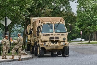National Guard soldiers standing next to a line of tan military trucks parked along a tree-lined street in Washington, DC.