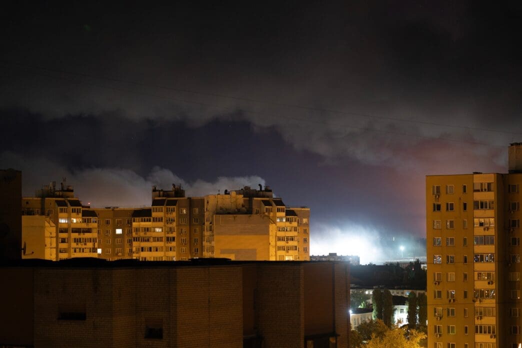A nighttime view of Kyiv apartment buildings, with a bright glow and smoke rising in the background from a Russian attack.