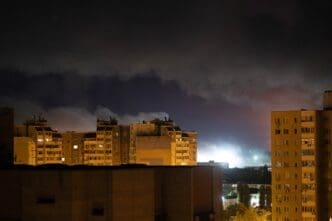 A nighttime view of Kyiv apartment buildings, with a bright glow and smoke rising in the background from a Russian attack.