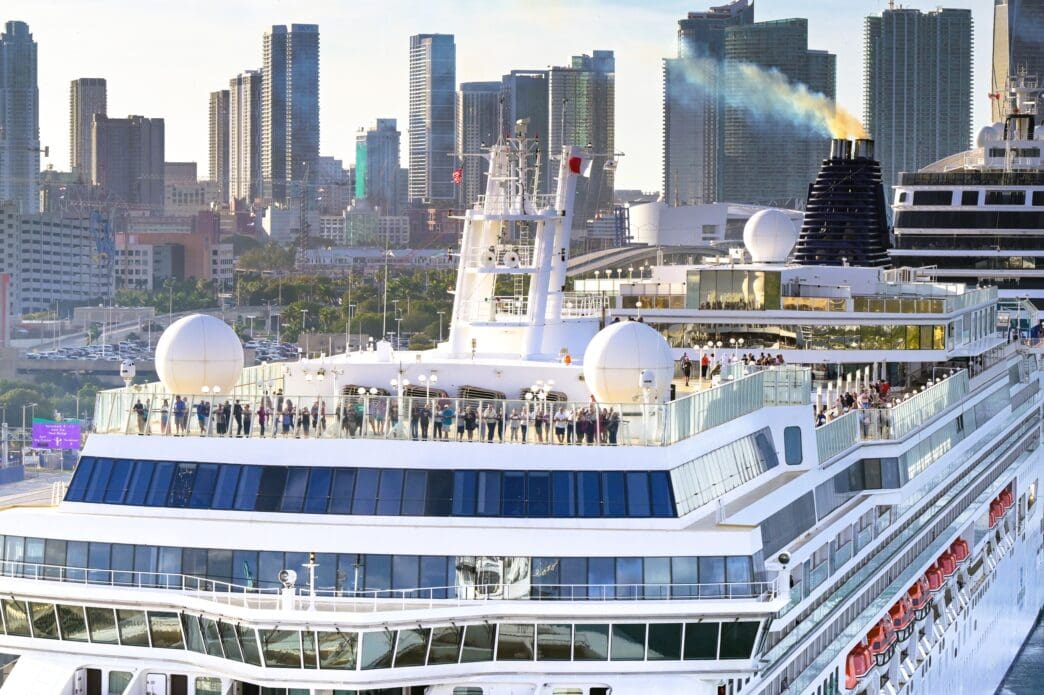 Passengers stand on the upper deck of the Norwegian Jade cruise ship, with smoke rising from its funnel against the Miami skyline.