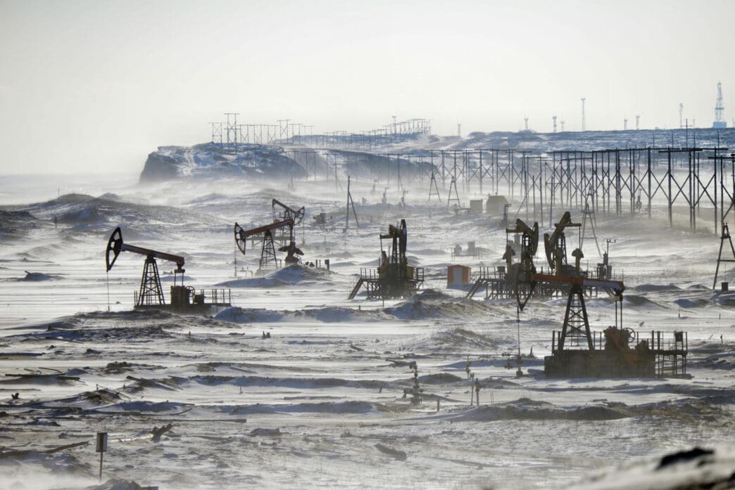 Numerous oil pumpjacks operate on a snow-covered landscape under a hazy sky